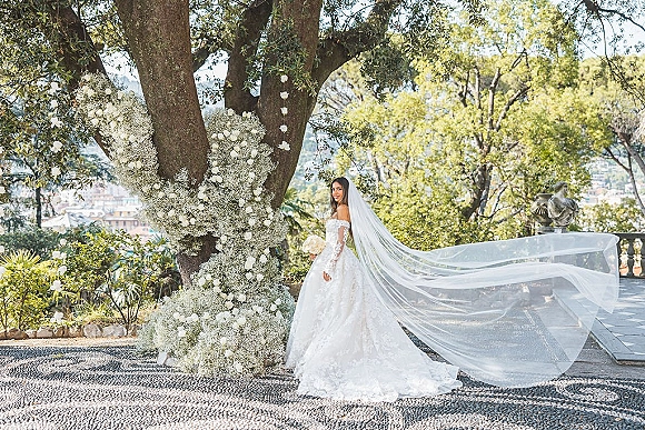 Bridal portrait of a bride with long veil in an off the shoulder lace wedding dress, holding a white rose bouquet on a garden terrace with city view