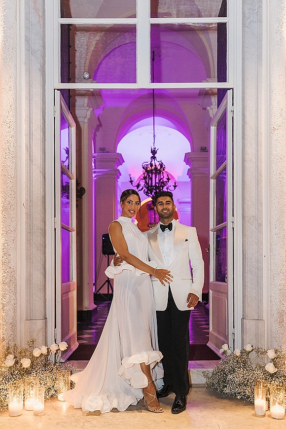 Couple portrait of bride and groom at a doorway, candlelit with white roses and baby's breath, beneath a chandelier and purple uplighting