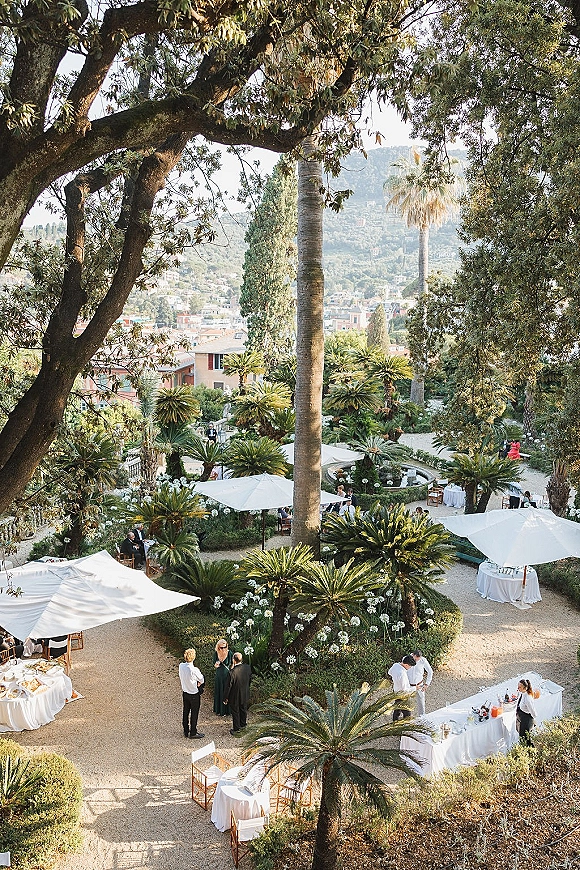 Outdoor wedding reception with white patio umbrellas, cocktail tables and bar setup with glassware, on a garden terrace with palm trees and mountain view