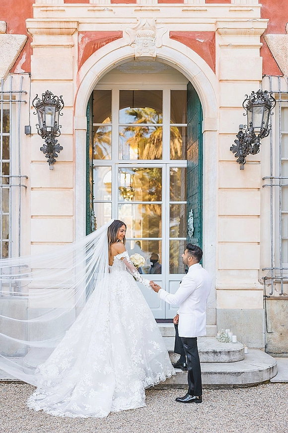 Couple portrait at an arched wedding doorway, bride in off-shoulder gown with long veil and bouquet holding hands with groom by lanterns and candles