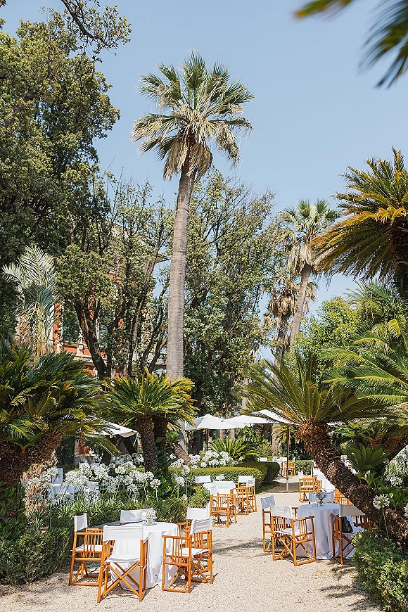 Outdoor reception setup with garden wedding reception seating, round tables under white umbrellas with floral centerpieces in a palm garden courtyard
