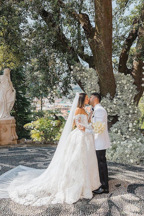 Wedding kiss portrait of bride and groom kissing under a baby’s breath floral arch, veil and white rose bouquet in cobblestone garden courtyard