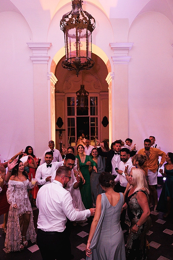 Wedding dance floor packed with wedding reception dancing guests in formal attire, cocktail glasses raised under chandelier and uplighting by arched doorway