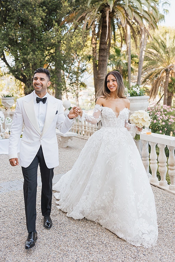 Couple portrait of bride and groom holding hands, bride with white rose bouquet in lace gown, groom in white tux by palm garden balustrade