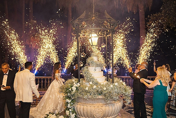 Wedding cake display with a tiered white cake and white rose topper, sparklers and hanging glass orbs on a string-lit terrace at night