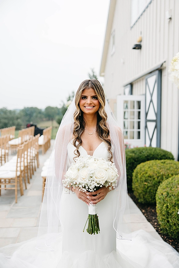 Bridal portrait of a bride holding bouquet with a cathedral veil, strapless gown, and white rose bouquet on an outdoor barn aisle