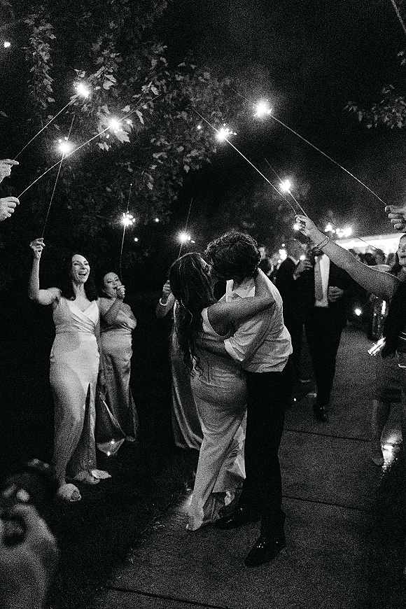 Wedding sparkler exit with a sparkler send off as bride and groom kiss in a glowing tunnel, guests lining an outdoor walkway under trees at night