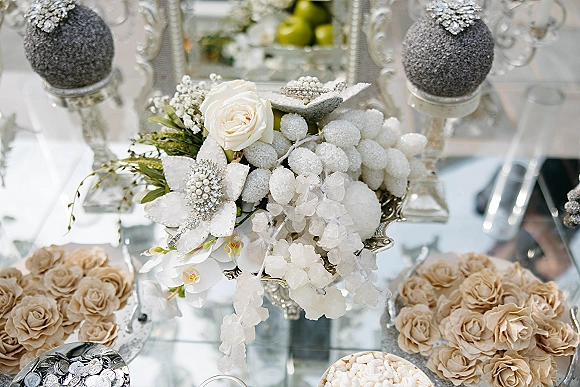 Wedding centerpieces with a white floral centerpiece of roses and orchids in a silver pedestal vase on a mirrored tabletop with glass candlesticks