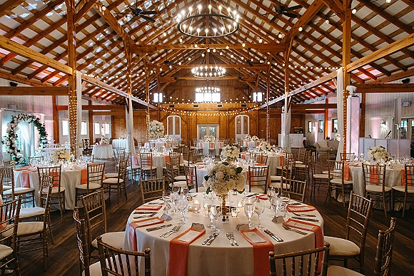 Wedding reception decor with round tables, coral runners, gold chiavari chairs, floral centerpieces and candles under string lights in a wood barn