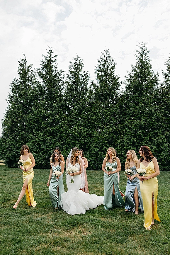 Bridesmaid group photo of bride with bridesmaids walking on a grass lawn, holding white rose bouquets in satin high-slit dresses under evergreens