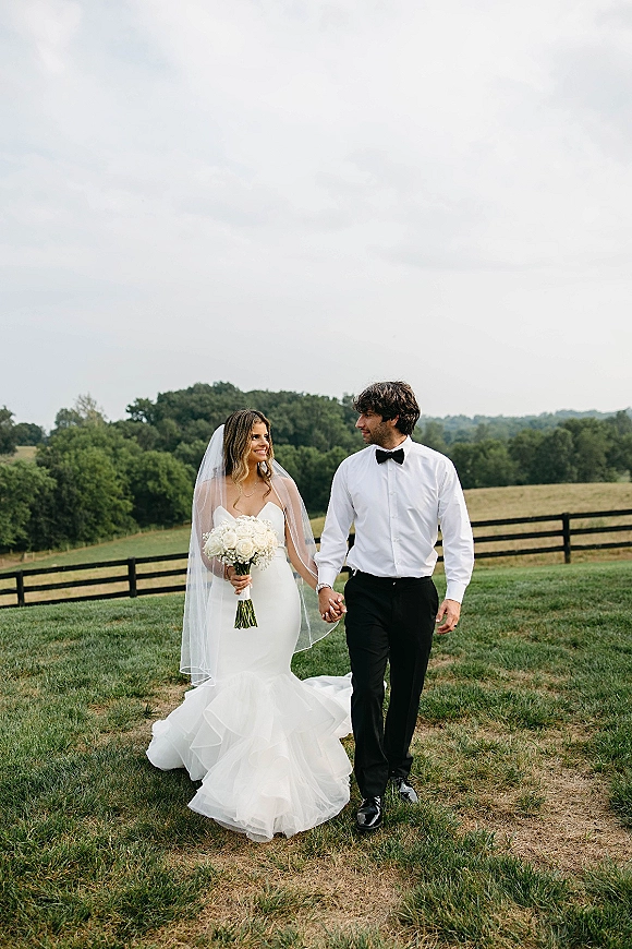 Couple portrait of bride and groom holding hands, she carries a white rose bouquet and long veil in a grassy field by a wooden fence