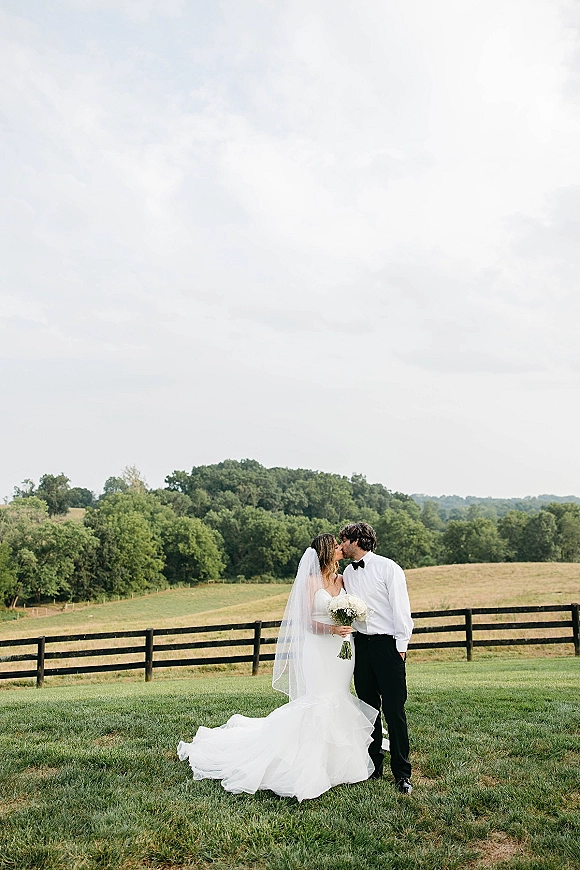 Wedding kiss portrait of bride and groom kissing, her veil and bouquet visible beside a wooden fence in a rolling pasture field