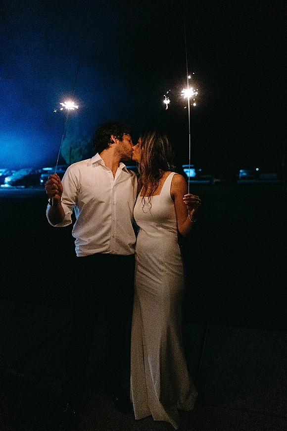 Wedding kiss portrait of newlyweds holding sparklers, bride in a wedding dress kissing groom in a white shirt under the night sky