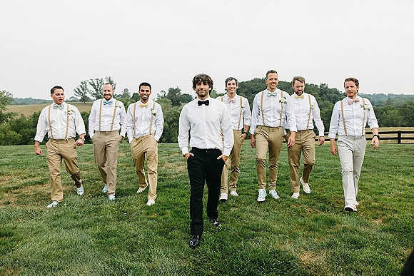 Groomsmen portrait with suspenders and bow ties, boutonnieres and mixed pants, walking in a grassy field by a split rail fence
