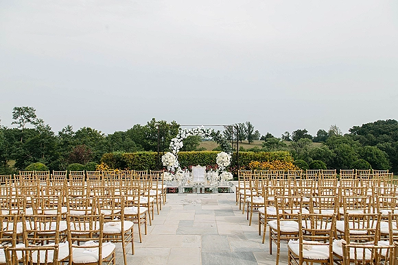 Ceremony setup with outdoor ceremony seating, gold Chiavari chairs and a glass aisle leading to a white floral arch on a stone patio garden