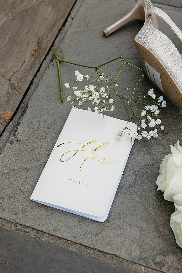 Wedding vows book in white with gold foil script beside bridal heels, baby's breath and white roses arranged on stone steps