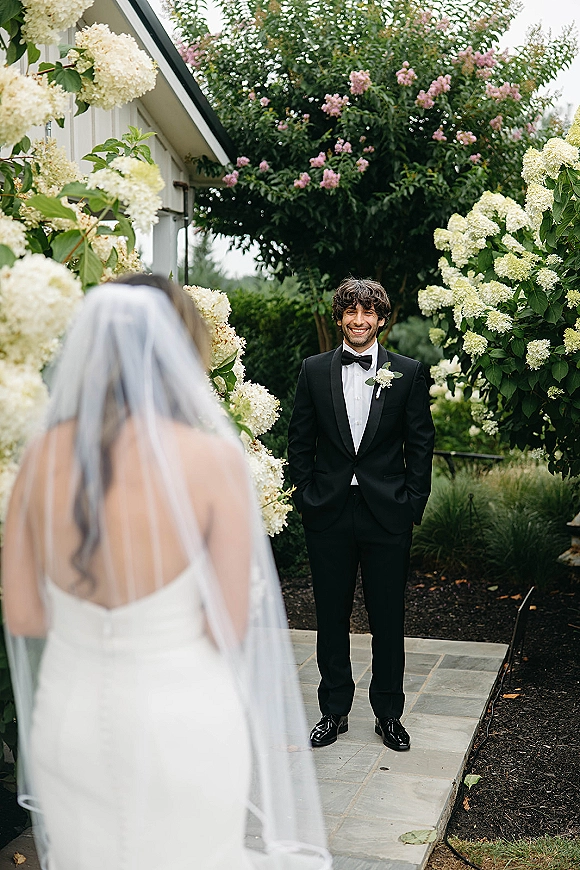 Wedding first look as groom first look reaction unfolds with the bride in a strapless dress and veil approaching him on a garden walkway