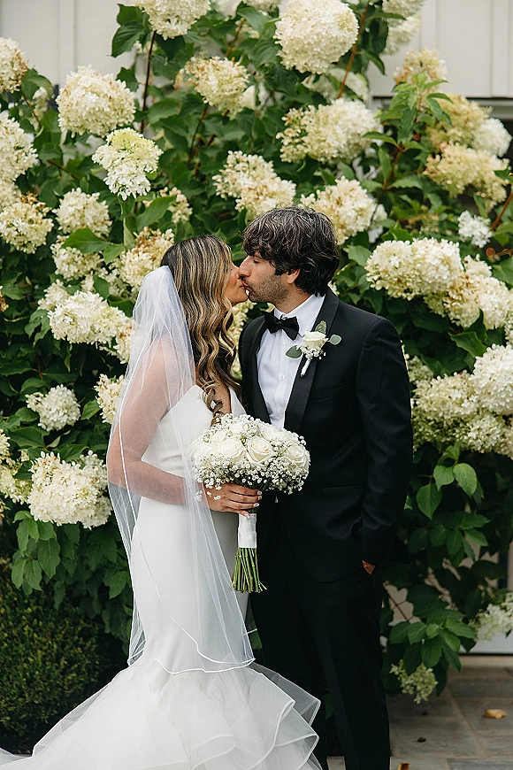 Wedding kiss portrait of bride and groom kissing, her veil and white rose bouquet with baby’s breath against hydrangea bushes outdoors