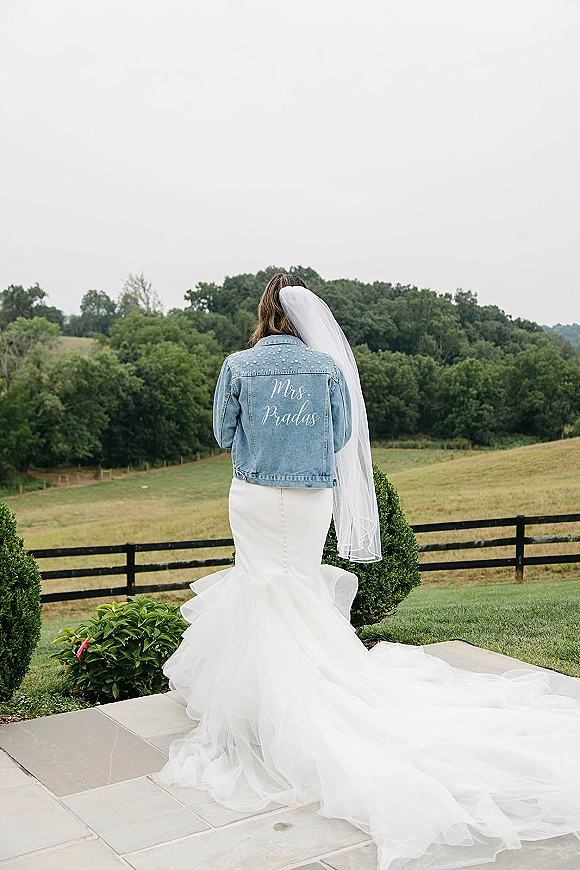 Bridal portrait of a bride from behind in a cathedral veil and long train, wearing a denim jacket on a stone patio by rolling hills