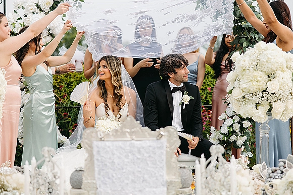 Wedding ceremony moment with bride and groom seated as bridesmaids lift her lace veil, surrounded by white florals and a garden arch