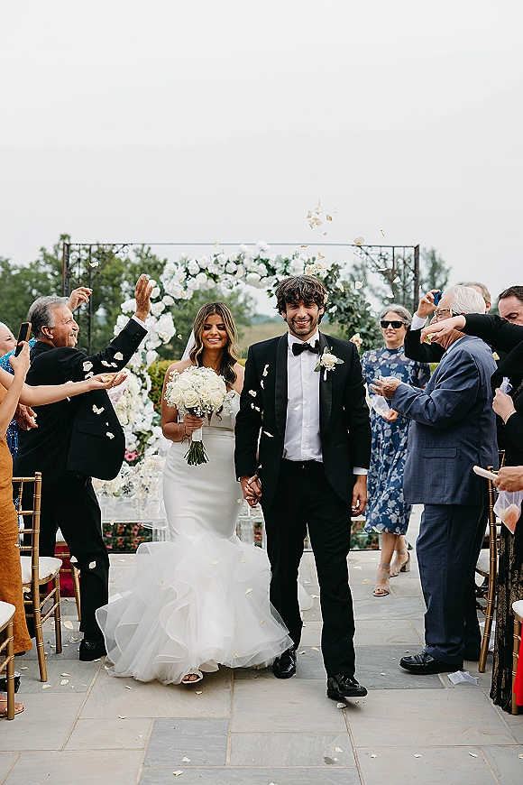 Wedding recessional as newlyweds walking down aisle holding hands, bride with white rose bouquet, rose petals flying under floral arch outdoors