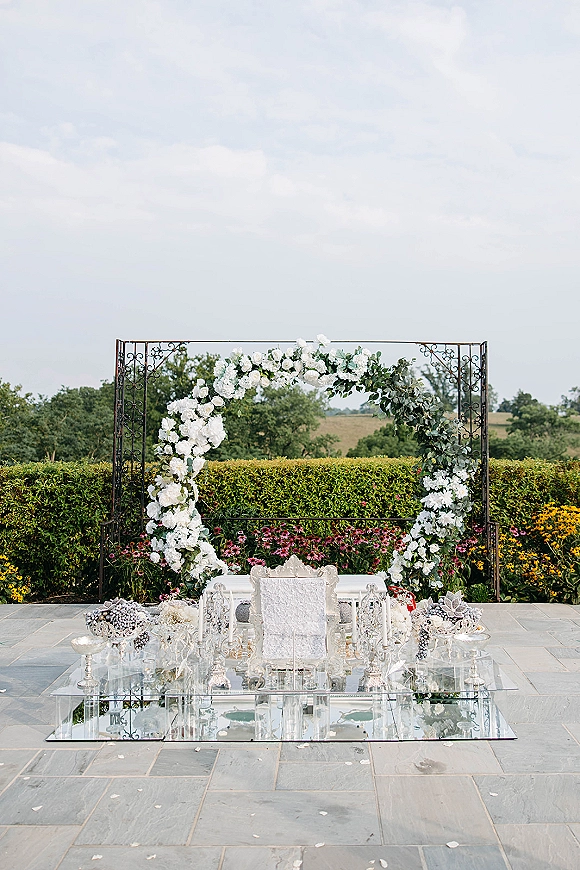 Wedding ceremony backdrop with a round floral arch of white blooms and greenery, mirrored aisle runner, and ornate chair on a garden patio