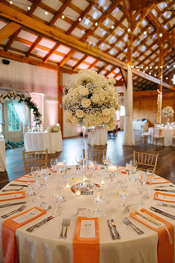 Reception tablescape with tall wedding centerpiece of white hydrangeas and roses in a glass trumpet vase on round table beneath string lights in a barn hall