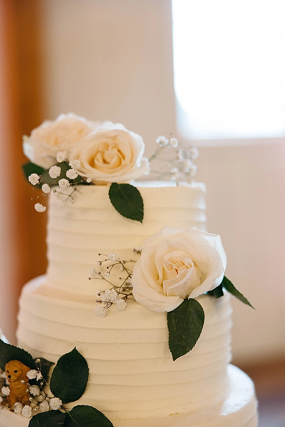 Wedding cake three tier wedding cake with white roses and baby's breath accents, buttercream frosting and greenery on a stand by window light