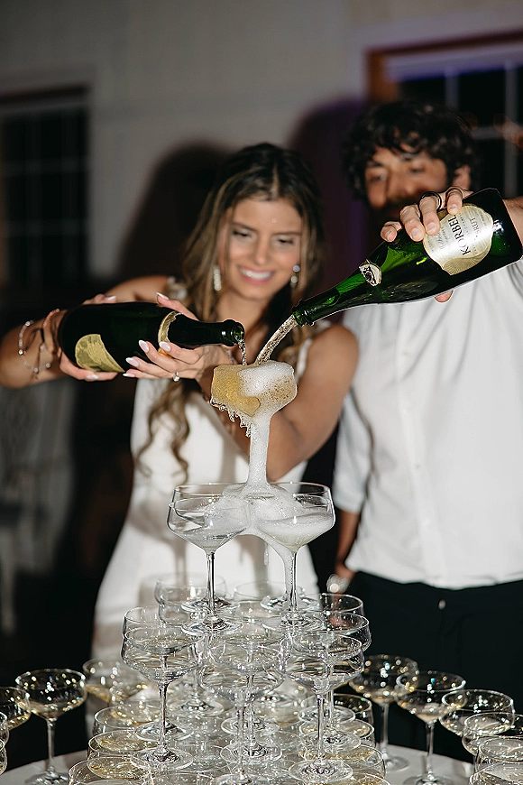 Champagne tower pour cascading through a coupe glass pyramid as bride and groom pour bottles indoors by a window, foam spilling over