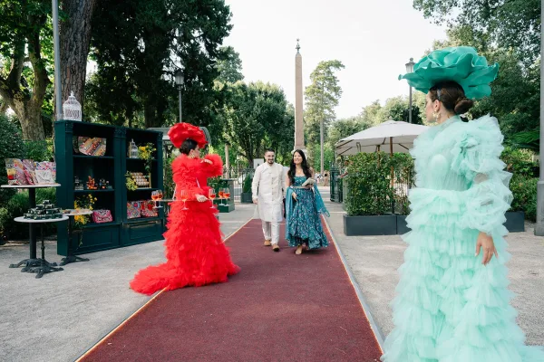 Couple entrance on a red carpet runner during wedding welcome drinks, approaching iron gates along a garden-lined paved walkway.