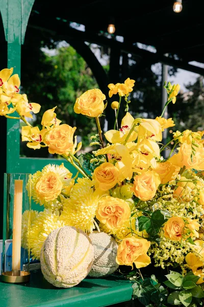 Wedding flower arrangement of yellow wedding flowers framing a glass hurricane candle, set against a green wooden structure with string lights.