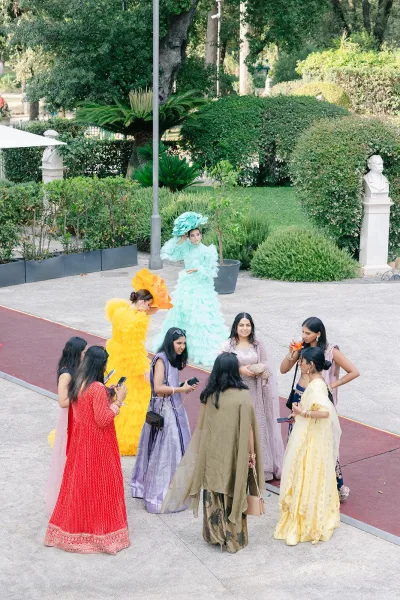 Wedding guests mingle at an outdoor cocktail hour, some in vibrant sarees, along a garden walkway bordered by hedges and a stone statue.