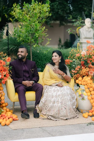 Couple portrait featuring a bride in lehenga and groom, set amid garden greenery with yellow armchairs adding a vibrant accent.