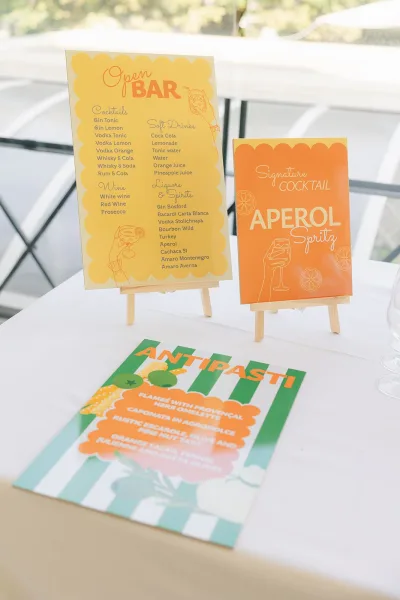 Wedding bar signage featuring a signature cocktail sign on an easel, displayed near a window with trees in the background.