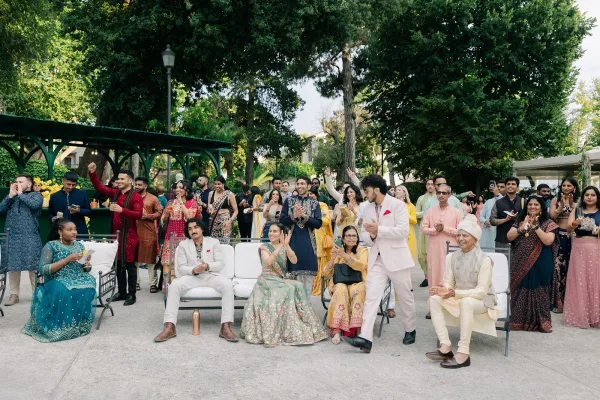 Wedding guests share a clapping moment in a garden under a pergola, some in vibrant saris, as sunlight filters through the trees.