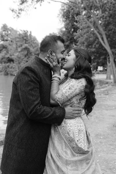 Wedding kiss portrait as a bride and groom kiss by a tree-lined shoreline, her dupatta catching light as they embrace beside calm water.