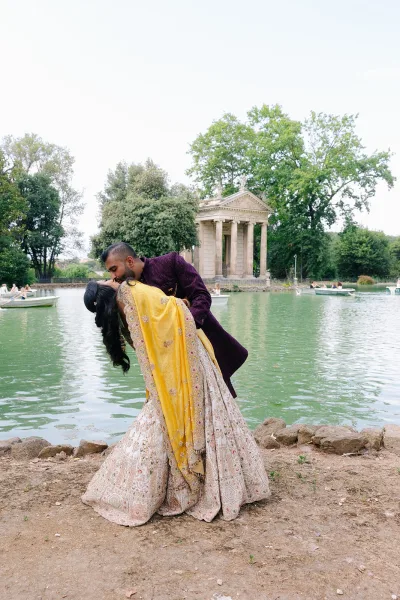 Wedding kiss portrait of a couple in a dip kiss pose lakeside, her yellow dupatta flowing, with a classical temple and rowboats behind.