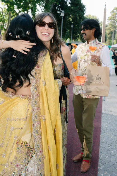 Wedding guest hug at an outdoor wedding cocktail hour, one in a bright saree, as they embrace beneath trees on a cobblestone walkway.