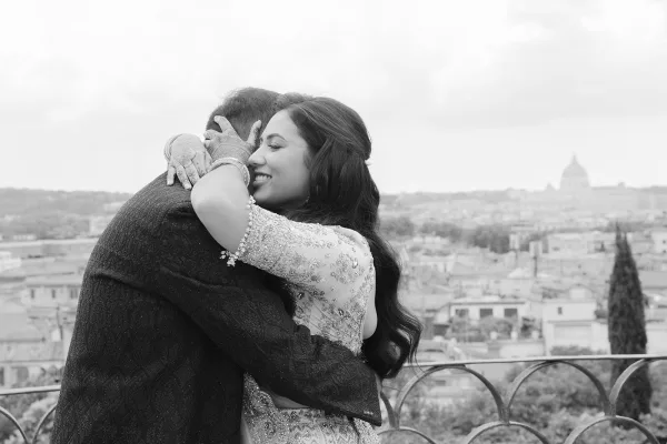 Wedding hug moment with a bride hugging groom on a balcony overlooking a city skyline and distant dome, her hands adorned with henna.