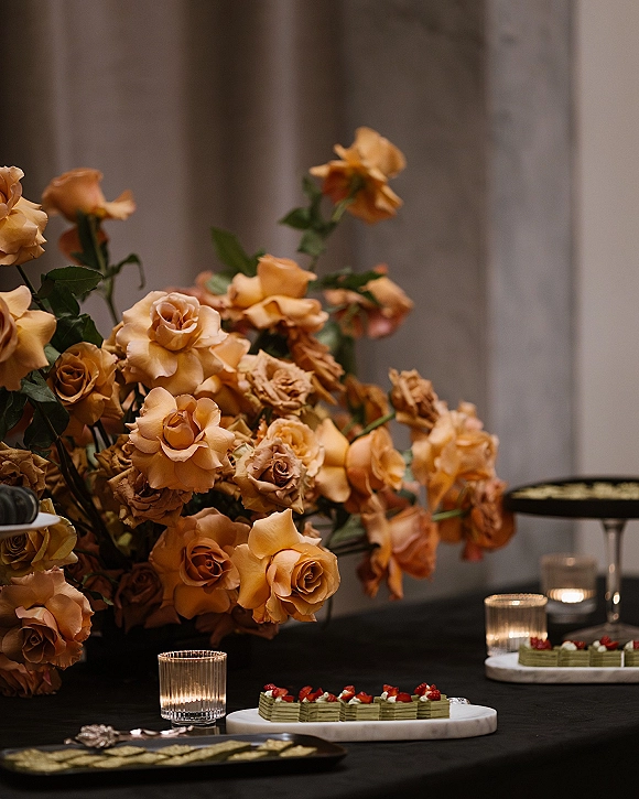 Wedding dessert table with petite wedding desserts, orange rose centerpiece and votive candles on black tablecloth against a stone wall indoors