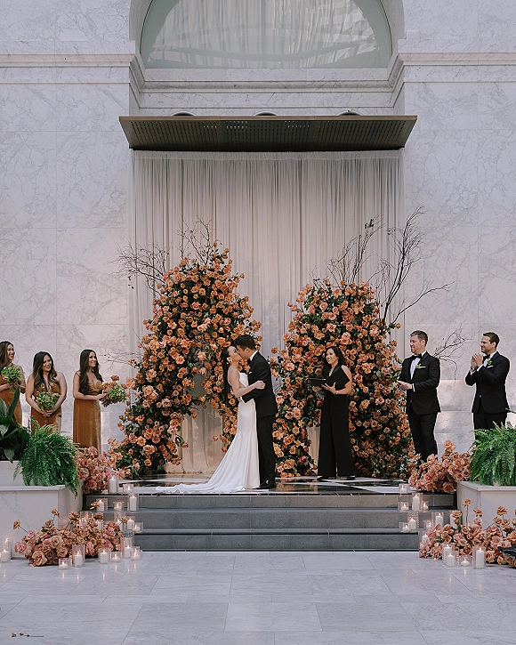 Ceremony kiss as bride in a strapless gown and groom in a black tuxedo embrace beneath a floral arch against marble wall and drapery