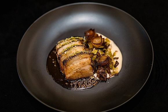 Wedding dinner plate with sliced meat, pan sauce, vegetable side and puree on a black ceramic plate against a dark table surface