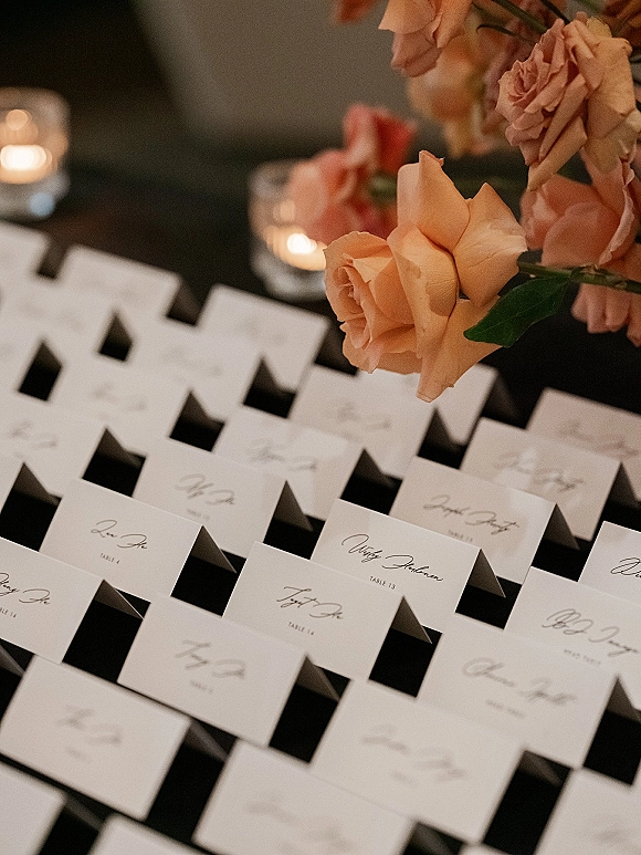 Wedding place cards with calligraphy place cards beside peach roses and glowing votive candles on a dark tabletop with soft blur behind