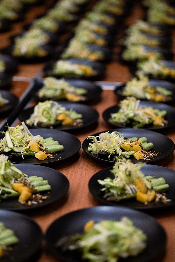 Wedding plated salad on a black dinner plate with greens, citrus segments, and seed topping, lined in rows on a banquet prep table