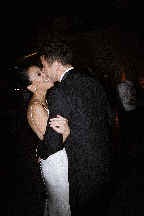 First dance as bride in a white wedding dress and pearl earrings smiles while groom in tuxedo holds her in a dark reception room