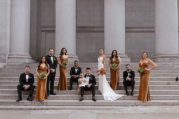 Wedding party portrait of bride, groom, bridesmaids and groomsmen posed on stone steps before tall columns, holding bouquets