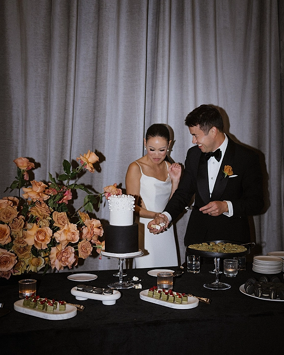 Wedding cake cutting as bride in satin gown and groom in black tux slice pearl cake at dessert table with candles, roses, gray drapery backdrop