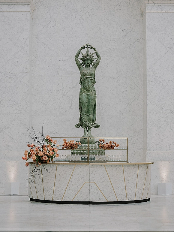 Wedding bar setup with a champagne bar wedding display of glassware, brass-trim marble counter, peach rose arrangement, and statue backdrop