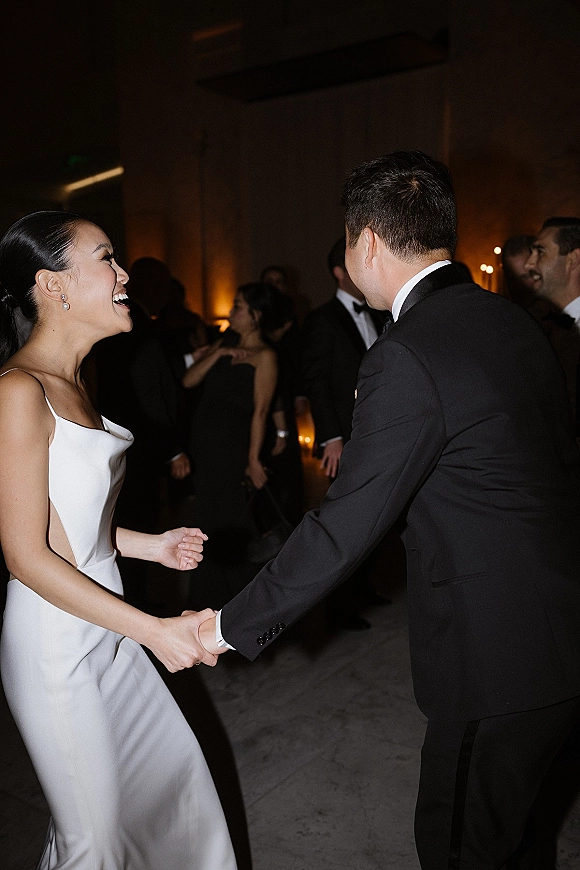 Wedding reception dancing as bride laughs holding groom’s hand, satin dress and pearl drop earrings amid guests in warm indoor lighting