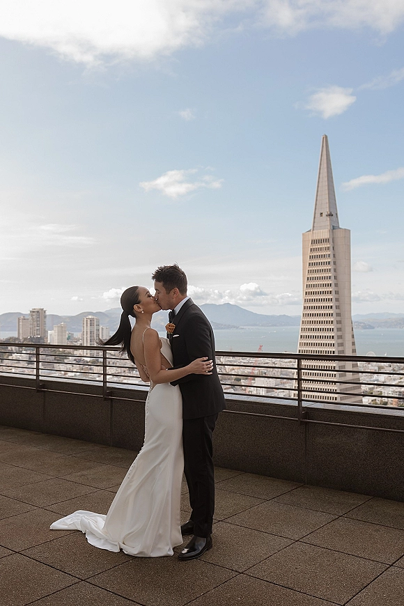 Wedding kiss portrait of bride in a simple slip dress and groom in black tuxedo embracing on a rooftop terrace with city skyline and bay beyond